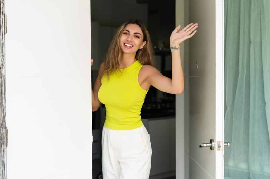 בחורה בגופייה צהובה מחייכת עומדת ומנופפת בכניסה לדלת